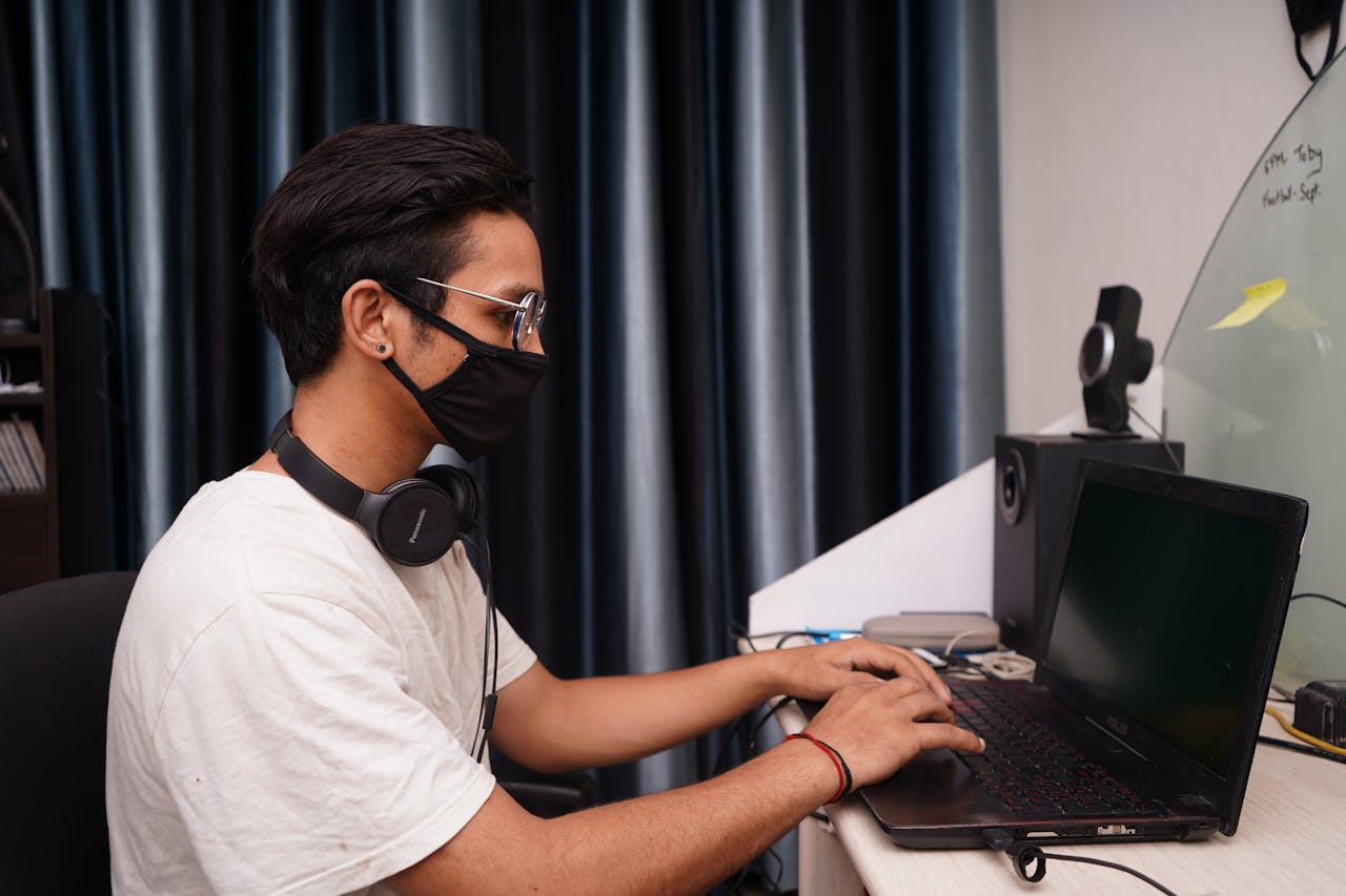 Crafting Captivating Headlines: Your awesome post title goes here A young man wearing a mask works on his laptop at a home office desk, surrounded by tech accessories.